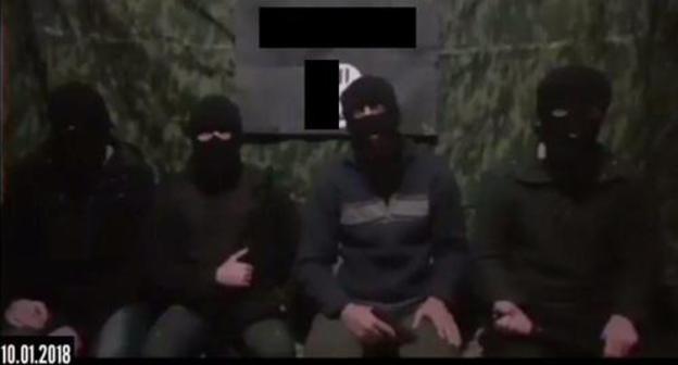 Four masked men sitting at the background of the IS'* flag call on their followers to kill Russian rights defenders. Screenshot of the video.