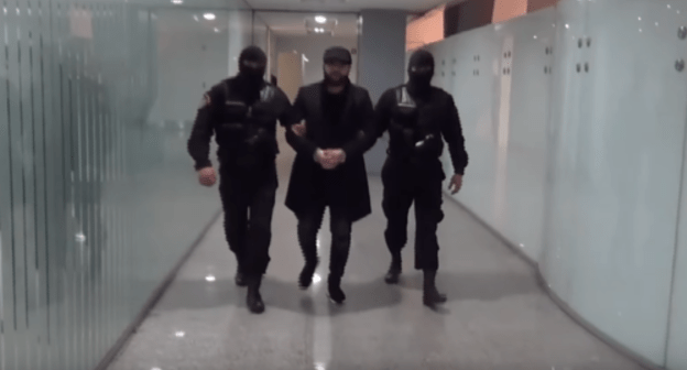 Two masked law enforcers are escorting handcuffed Narek Sargsyan at Zvartnots Airport in Yerevan. Screenshot from video posted at: https://www.youtube.com/watch?time_continue=16&v=1MUWqxKFicE&feature=emb_title